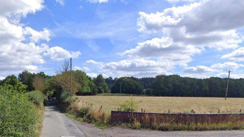A narrow country road with large field to the right with trees and bushes surrounding on either side.
