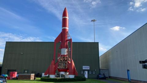 A 60ft tall red, Thunderbird rocket. It's on a grass field in front of a green building. It has white accent stripes