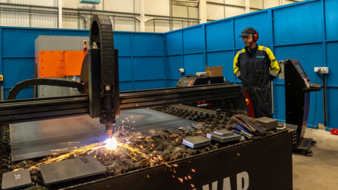 A man in high vis protection gear, including a cap and ear defenders, stood in front of a factory line where metal is being shapes by a machine and sparks are flying off it.