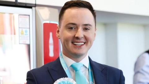 Reform deputy leader Darren Grimes. He has short brown hair that are shorter on the sides and combed to one side. He has green eyes and is smiling. He wears a navy suit, with light blue tie, lanyard and rosette. The background is blurred.