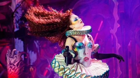 Chappell Roan performs onstage. Her long red hair is splayed out behind her as she moves across the stage. Her Tudor-esque burlesque outfit has her face on the front.