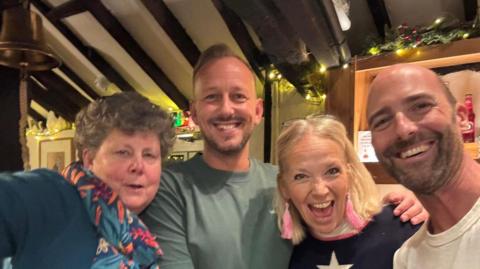 A lady with short curly hair wearing a blue and red scarf (pub volunteer Geraldine Symonds) pictured smiling with three other local members of the community - two men and a woman. They are standing in the Swan Pub with Christmas fairy lights and the pub bell in the background. 