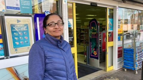 A woman with her hair tied back, wearing glasses and a blue puffer coat standing in front of the open doors of a shop