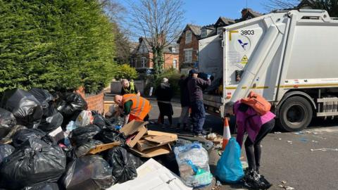 The back of a bin lorry on a residential road is being filled with piles of rubbish that are mounted up on the floor. Several people are helping to move the rubbish from the floor into the truck.
