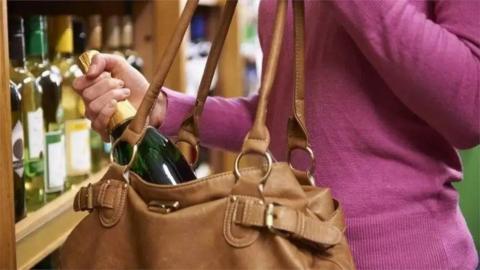 A woman is wearing a pink jumper and is holding a brown handbag. She is standing next to a shelf of wine and placing a bottle into her handbag. 