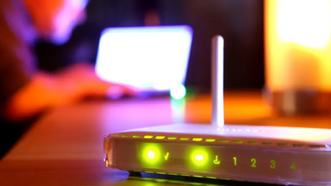 A white broadband route is lit up with green light in front of a blurry background of a person working on a laptop.