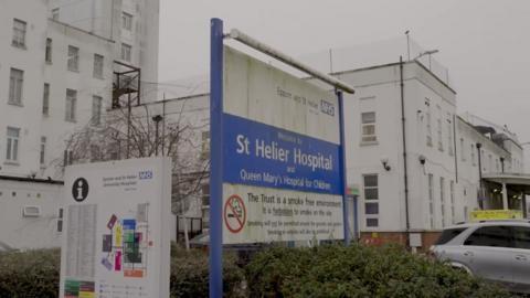 The St Helier Hospital sign outside the hospital, looking dirty, with the wording 'St Helier Hospital and Queen Mary's Hospital for Children'. The large board is blue and white. 