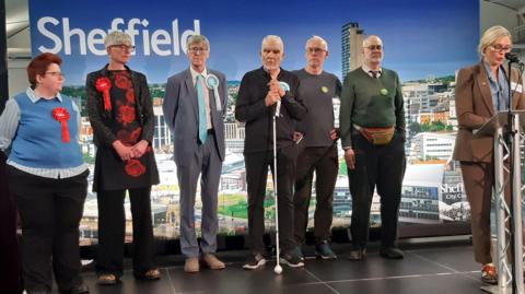 A group of seven people standing on a stage.

The stage is set in front of a large printed backdrop featuring a city skyline and the word "Sheffield" in large white letters across the top left. In the lower right corner of the backdrop, the text "Sheffield City Council" is visible alongside a city crest or logo.

Six people stand in a line across the stage while a seventh person stands at a lectern on the far right. Several members of the group are wearing coloured rosette badges attached to their clothing. Two red rosettes are visible on the people standing on the left side of the stage, while blue and green rosettes can be seen on some of the people standing toward the centre and right.