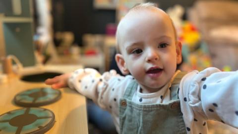 Wren standing up against a wooden table, wearing a green pinafore and white shirt with black dots