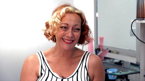 A woman with short blonde hair wearing a black and white stripped top sitting in front of a mirror
