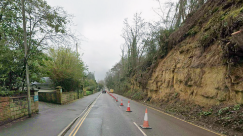 A road is seen running alongside a bank with trees on top. One side of the road is closed.