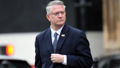 Close up of Andrew Rosindell walking outside wearing a suit with a union jack flag pin on the lapel . He is looking to his left