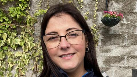 Shows a young woman with dark long hair and glasses smiling. She is standing in front of a wall, with ivy and a hanging flower pot to the right of the picture. 