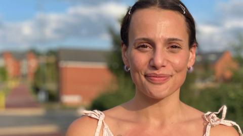 A head and shoulders shot of Saffron McCubbin who is smiling and looking at the camera. She is wearing a cream top. The background behind is blurred, but it shows red brick homes, a road, some yellow bollards and some bushes. 