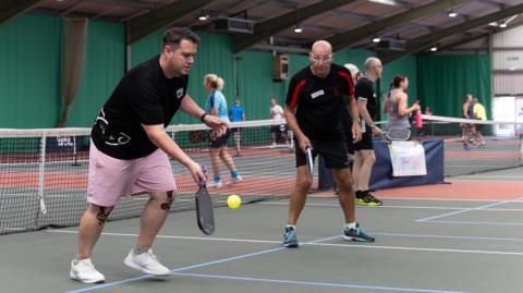 Mike Brockwell playing pickleball with a friend on an indoor court. There is a net behind them and other games taking place in the background.