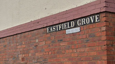 A red brick wall with a black and white sign plaque saying "EASTFIELD GROVE" 