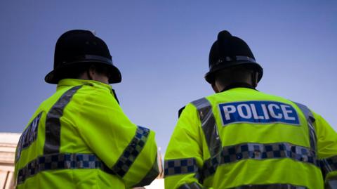 Two male police officers stand together with their backs to the camera. They are wearing high-visibility jackets. 
