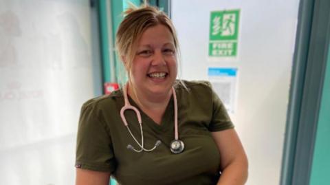 Claire in a midshot sitting on a stool at a high table in a clinic-type room in front of a fire exit door. She has a pink stethoscope round her neck and is wearing a green t-shirt. Her blonde hair is scooped up behind in a clip and she is smiling with her fingers interlaced.