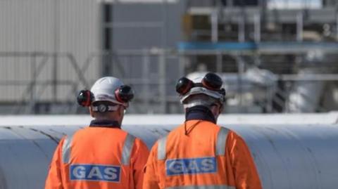 Two workmen in orange hi-vis jackets with white helmets on are walking in front of a large pipe and industrial site.