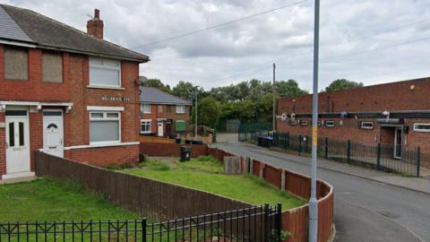 A street view of Millbrook Avenue. Semi-detached houses with line the street to the left. The street sign is fixed to one of the houses.