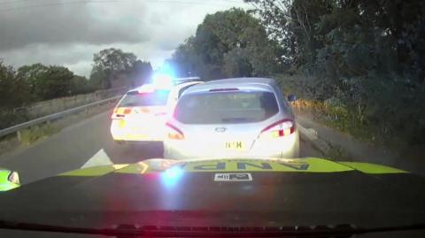 A silver Ford Mondeo with a police car with blue light to its left and one behind, boxing it in
