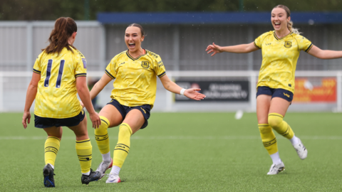 Three female footballers all wearing a yellow and dark navy blur football kit seemingly celebrating a goal together on a football pitch.