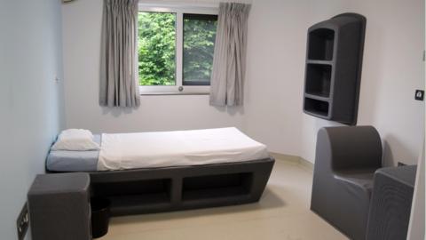 A room at the new ward, with a single bed under a window, soft furnishings and a plastic shelving unit