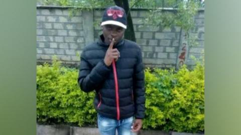 Boniface Kariuki in a black jacket and cap, holding his finger to his lips