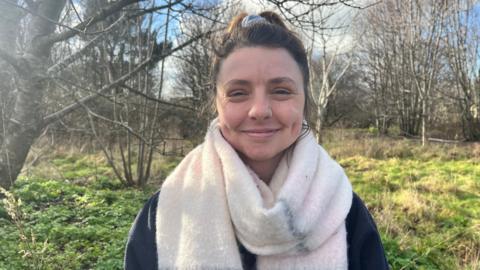 A woman smiling into the camera. She is wearing a thick, white scarf with a navy jumper and her brown hair is tied back in a scrunchie. She is stood in a woodland, and there is green plants behind her, along with bare trees. 