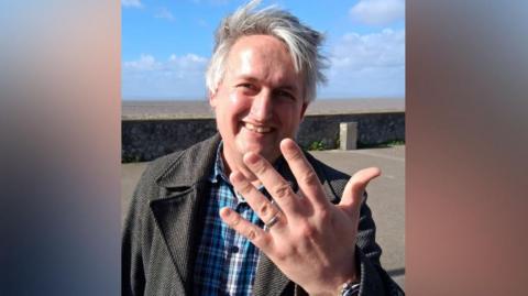 A man with light-grey hair and a blue checked shirt is smiling and holding out his hand which has a silver wedding band on it.