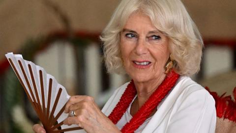 Queen Camilla at the Commonwealth summit in Samoa on 24 October, she is smiling slightly and has a gold-coloured chair behind her, and holds a red and white hand fan in her hands