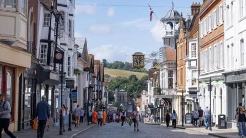 An view down a small high street lined with historic buildings. In the background are green hills and trees