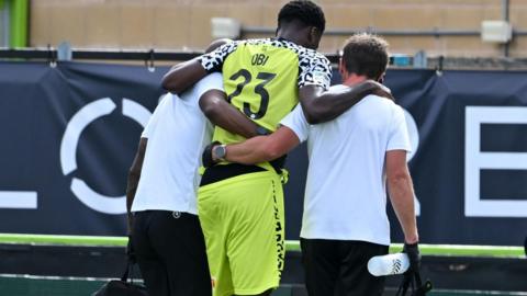 Emeka Obi in a pale yellow shirt and shorts being helped off by two men in white shirts who have their arms round him