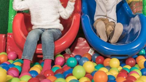 A stock image of two children on a slide each with their feet in a ball pool. Their faces are not in shot.