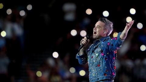 Robbie Williams performs on stage in a denim jacket with colourful gems stuck all over it. He has one hand in the air with his palm up to the sky and is singing into the microphone. The crowd is blurred with circular lights visible in the background.