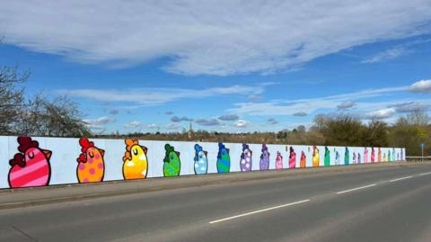 A mural on a wall at the side of a road shows paintings of chickens in various colours