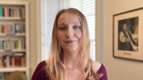 A girl in her 20's with blonde hair and blue eyes smiles at the camera. She is wearing a purple top and necklace. In the background is a framed picture and bookcase.