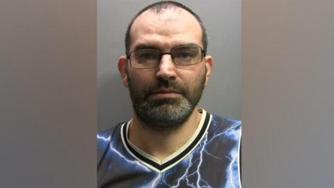 A mugshot of Raif Mentesh. He has balding dark hair which is cut short and stubble with flecks of grey hair. He has black rectangular glasses and wears a purple basketball vest which has lightning bolts on it. 