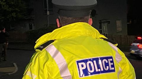 A male police officer wearing a hi-vis jacket that reads police. He has his back towards the camera