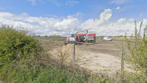 A picture of one of the fields where the solar farm will be built. A metal fence has been erected around the field next to a row of hedges. A digger is parked in the field near to a white van and a pile of earth.