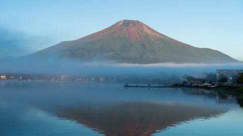 A wide shot of Mount Fuji seen without snow, silhouetted in a blue lake, in September last year