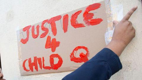 A hand places a sign on a wall reading "Justice 4 Child Q" in red paint.