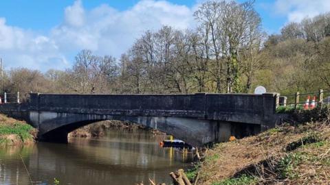 The picture shows a low, concrete road bridge crossing a calm river in a rural setting. The bridge has a single wide arch and darkened concrete, with visible staining along the sides that suggests age and weathering. Metal railings run along the top edges of the bridge, and there are a few orange-and-white traffic cones placed near one end.