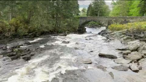 A rocky treelined river crossed by an ancient stone footbridge