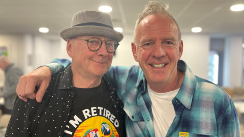 Norman cook wearing a blue chequered shirt smiling with his uncle Dennis whos wearing a black shirt and grey cap