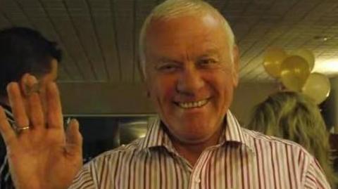 Frank Knight has white hair and a white shirt with coloured stripes