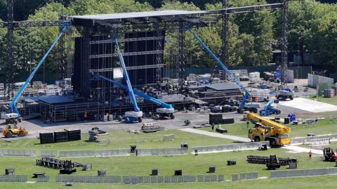 Cranes and other heavy machinery is used to set up a large stage in a wide, open park. 