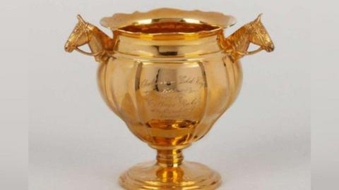 A solid gold cup trophy from the 1940s. The cup is quite bulbous, and the handles are two horse's heads, wearing bridles. The rim of the trophy is fluted.