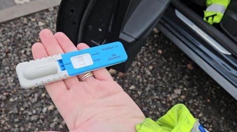 A police officer's hand holding a blue and grey drug testing kit.
