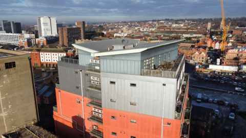 An aerial view of Cardinal Lofts, a tower block. The lower storeys that are visible have orange cladding, the upper floors lighter, grey cladding. The building is next to other tall buildings. Ipswich is in the background.
